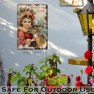 Puede incluir: Un letrero de metal de estilo vintage con una ni&ntilde;a peque&ntilde;a con un abrigo rojo sosteniendo un cervatillo. El letrero dice "Peace on Earth" y tiene un aspecto desgastado y r&uacute;stico. El letrero es perfecto para agregar un toque de encanto vintage a la decoraci&oacute;n de tu hogar.