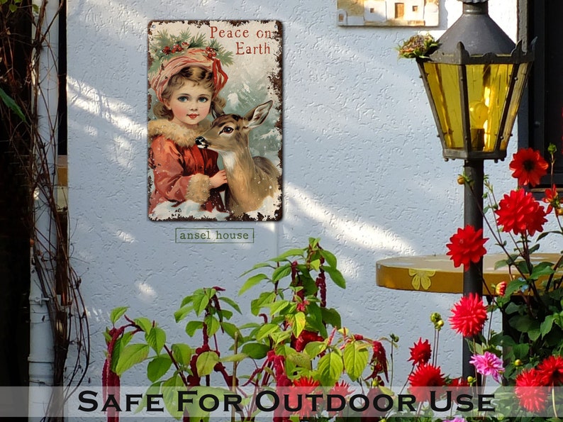 Puede incluir: Un letrero de metal de estilo vintage con una ni&ntilde;a peque&ntilde;a con un abrigo rojo sosteniendo un cervatillo. El letrero dice "Peace on Earth" y tiene un aspecto desgastado y r&uacute;stico. El letrero es perfecto para agregar un toque de encanto vintage a la decoraci&oacute;n de tu hogar.