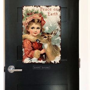 Puede incluir: Un cartel navide&ntilde;o vintage con una ni&ntilde;a joven con un vestido rojo y un cuello de piel blanca sosteniendo un cervatillo. El cartel tiene un aspecto desgastado y oxidado y dice "Peace on Earth".