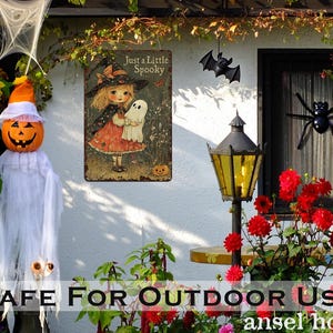 May include: Halloween-themed outdoor decorations. A pumpkin-headed figure in a white sheet, a metal sign with a witch and ghost, and a black bat and spider. The sign reads "Just a Little Spooky."