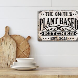 Op de afbeelding: Een wit bord met zwarte tekst dat "The Smith's Plant Based Kitchen Est. 2021" zegt. Het bord heeft een verweerde uitstraling en hangt aan een witte muur.