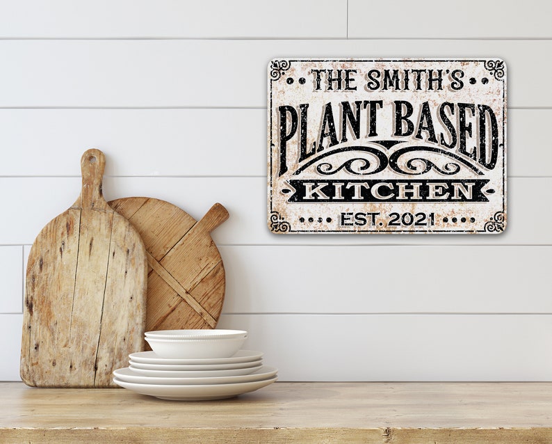 Op de afbeelding: Een wit bord met zwarte tekst dat "The Smith's Plant Based Kitchen Est. 2021" zegt. Het bord heeft een verweerde uitstraling en hangt aan een witte muur.
