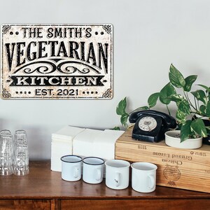 May include: A vintage-style metal sign with a distressed white background and black text. The sign reads "The Smith's Vegetarian Kitchen Est. 2021".