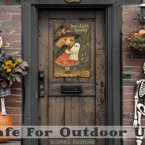 May include: A vintage-style Halloween sign with a witch and ghost illustration hangs on a wooden door. The sign reads "Just a Little Spooky." A scarecrow, pumpkins, and a skeleton decorate the porch, creating a festive autumn scene.