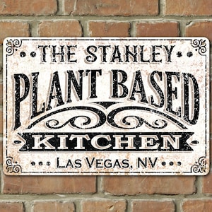 Op de afbeelding: Een wit metalen bord met zwarte tekst die "The Stanley Plant Based Kitchen... Las Vegas, NV..." luidt. Het bord heeft een verweerde, vintage uitstraling.