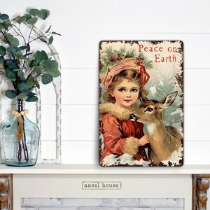 Puede incluir: Un letrero de metal de estilo vintage con un acabado desgastado que presenta a una ni&ntilde;a con un abrigo rojo sosteniendo un cervatillo. El letrero dice "Peace on Earth" en letras rojas.