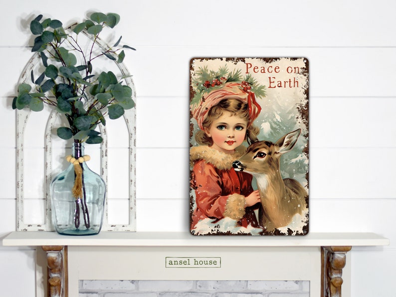 Puede incluir: Un letrero de metal de estilo vintage con un acabado desgastado que presenta a una ni&ntilde;a con un abrigo rojo sosteniendo un cervatillo. El letrero dice "Peace on Earth" en letras rojas.