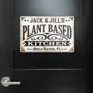 Op de afbeelding: Een wit bord met zwarte tekst dat "...Jack & Jill's... Plantaardige keuken... Boca Raton, FL..." zegt. Het bord heeft een verweerde, vintage uitstraling.