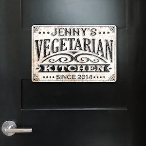 May include: A white metal sign with black text that reads "...Jenny's...Vegetarian Kitchen...Since 2014..." The sign has a distressed, vintage look.