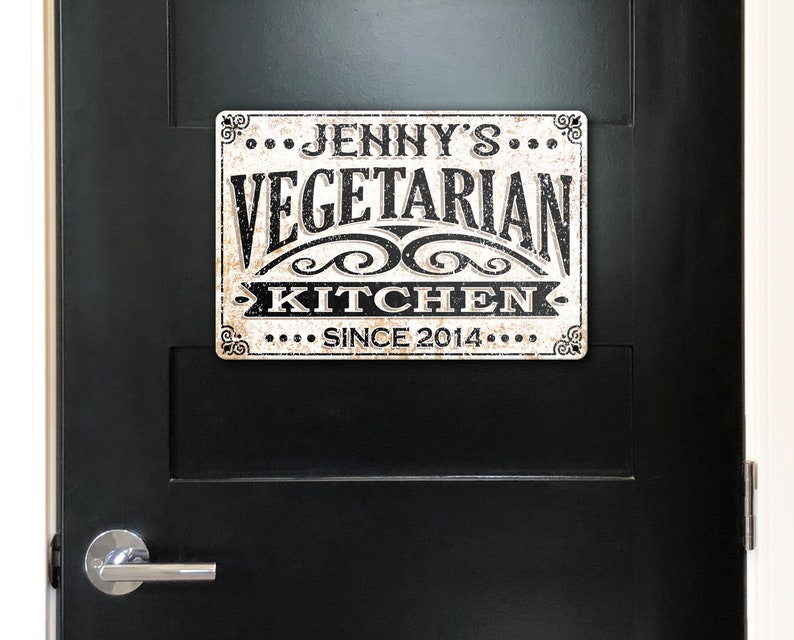 May include: A white metal sign with black text that reads "...Jenny's...Vegetarian Kitchen...Since 2014..." The sign has a distressed, vintage look.