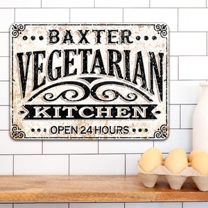 May include: A white metal sign with black text that reads "... Baxter ... Vegetarian Kitchen ... Open 24 Hours ..." The sign has a distressed look and is hanging on a white tiled wall.