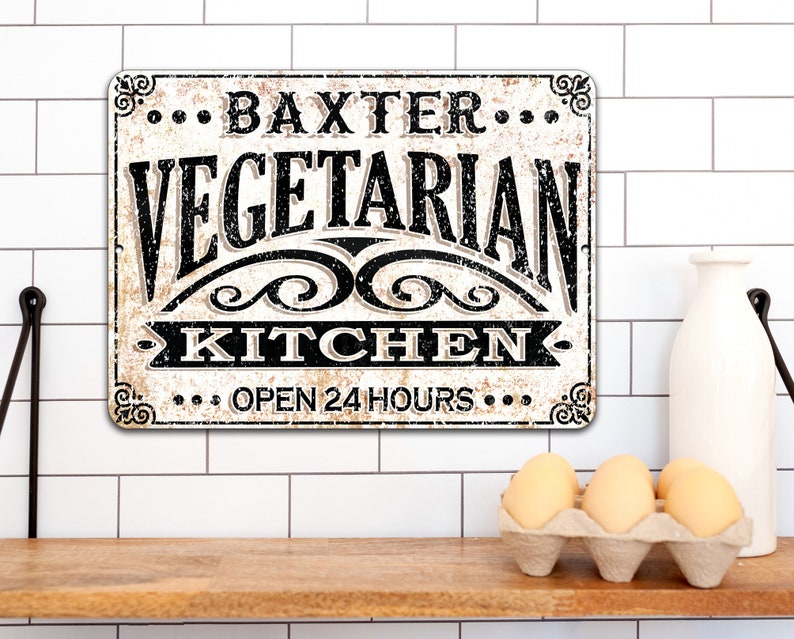 May include: A white metal sign with black text that reads "... Baxter ... Vegetarian Kitchen ... Open 24 Hours ..." The sign has a distressed look and is hanging on a white tiled wall.