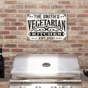 May include: A white metal sign with black text that reads "The Smith's Vegetarian Kitchen Est. 2021" hanging on a brick wall above a stainless steel grill.