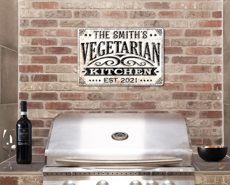May include: A white metal sign with black text that reads "The Smith's Vegetarian Kitchen Est. 2021" hanging on a brick wall above a stainless steel grill.