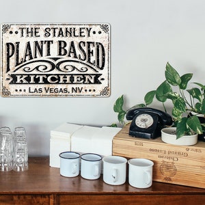 Op de afbeelding: Een vintage metalen bord met een verweerde witte achtergrond en zwarte tekst die "The Stanley Plant Based Kitchen Las Vegas, NV" luidt.