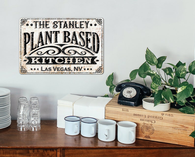 Op de afbeelding: Een vintage metalen bord met een verweerde witte achtergrond en zwarte tekst die "The Stanley Plant Based Kitchen Las Vegas, NV" luidt.