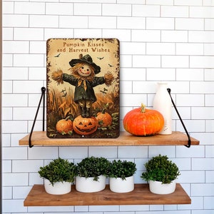 May include: A rectangular metal sign with the text "Pumpkin Kisses and Harvest Wishes" features a scarecrow standing on a carved pumpkin. An orange pumpkin and a white bottle sit on a wooden shelf. Below, are four white pots with green plants.
