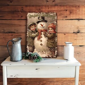 May include: A vintage-style Christmas sign featuring a snowman with two children. The sign has a red, white, and green color scheme and says "Seasons Greetings".