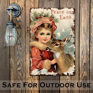 Puede incluir: Un letrero de metal de estilo vintage con una ni&ntilde;a con un abrigo rojo y un sombrero blanco sosteniendo un cervatillo. El letrero tiene un aspecto desgastado y dice "Peace on Earth".