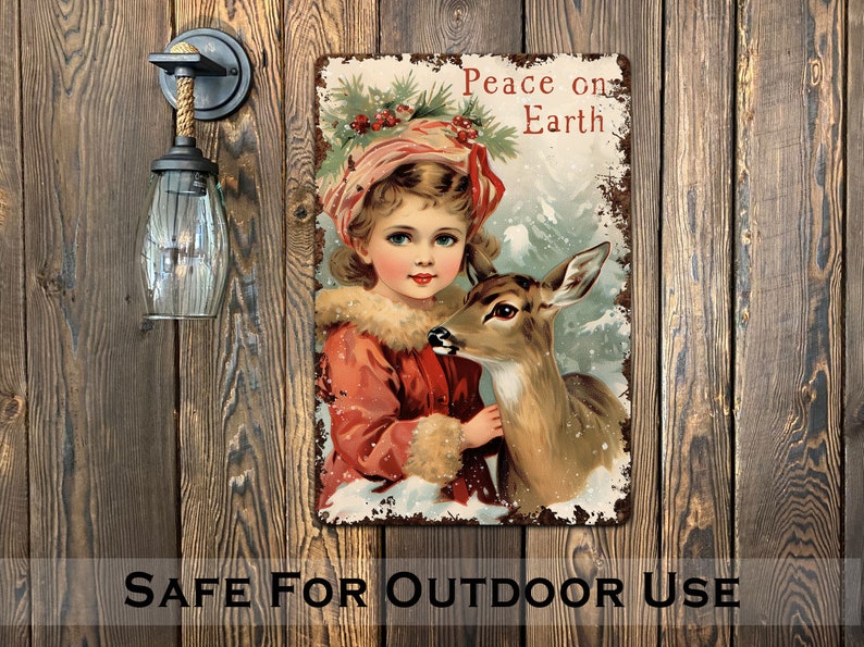Puede incluir: Un letrero de metal de estilo vintage con una ni&ntilde;a con un abrigo rojo y un sombrero blanco sosteniendo un cervatillo. El letrero tiene un aspecto desgastado y dice "Peace on Earth".