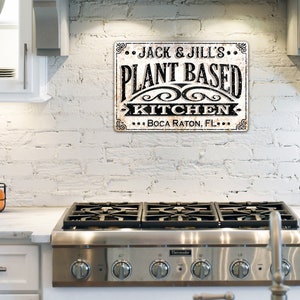 Op de afbeelding: Een witte bakstenen muur met een zwart-wit bord waarop staat "... JACK & JILL'S ... PLANT BASED KITCHEN ... BOCA RATON, FL ..." Een fornuis van roestvrij staal met vier branders staat op de voorgrond.