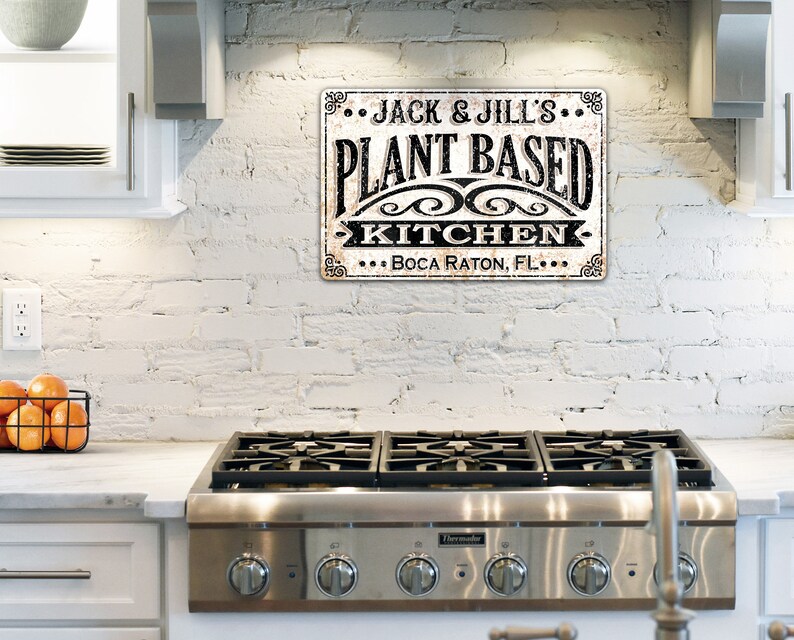 Op de afbeelding: Een witte bakstenen muur met een zwart-wit bord waarop staat "... JACK & JILL'S ... PLANT BASED KITCHEN ... BOCA RATON, FL ..." Een fornuis van roestvrij staal met vier branders staat op de voorgrond.