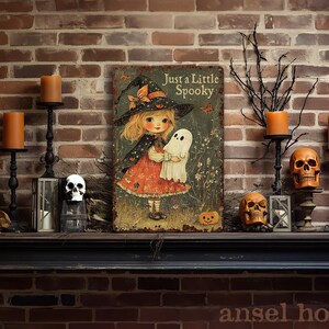 May include: A Halloween-themed metal sign with the text "Just a Little Spooky" featuring a witch holding a ghost. The sign is surrounded by candles, skulls, and a pumpkin on a brick mantel.