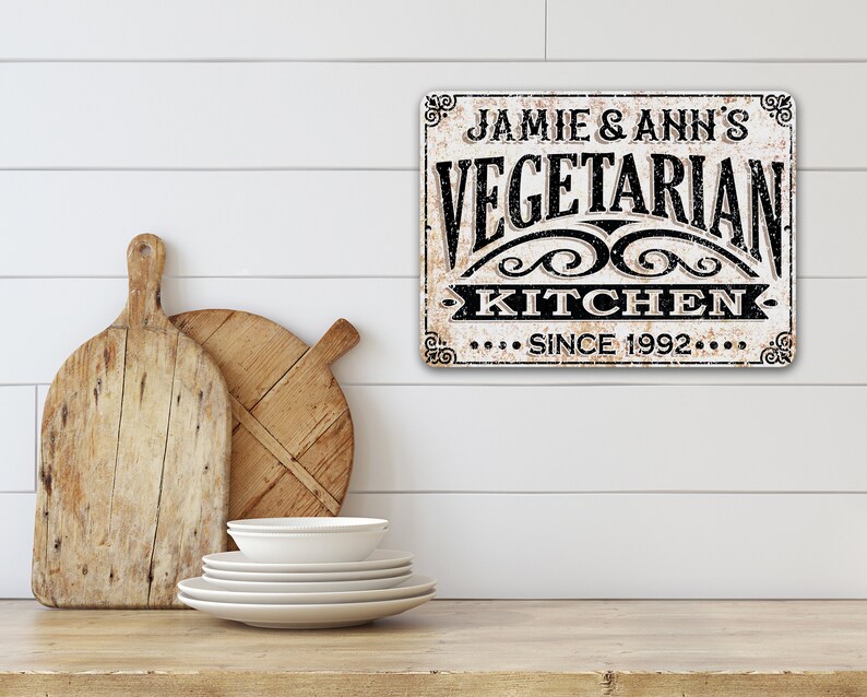 May include: A white sign with black text that reads "Jamie & Ann's Vegetarian Kitchen Since 1992". The sign has a distressed, vintage look and is decorated with ornate flourishes.