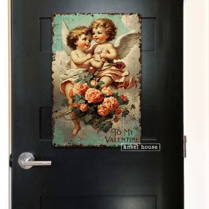 May include: Vintage-style metal sign featuring two cherubs with wings, holding a bouquet of pink roses. The sign has a distressed, rusty look and reads "To My Valentine" with the text "ansel house" below.