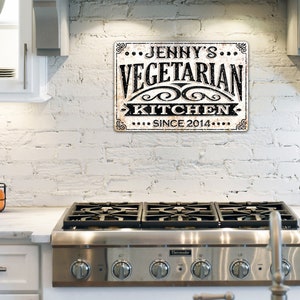 May include: A white brick wall with a black and white sign that reads "...Jenny's...Vegetarian Kitchen...Since 2014..."  The sign is hanging above a stainless steel stove with four burners.
