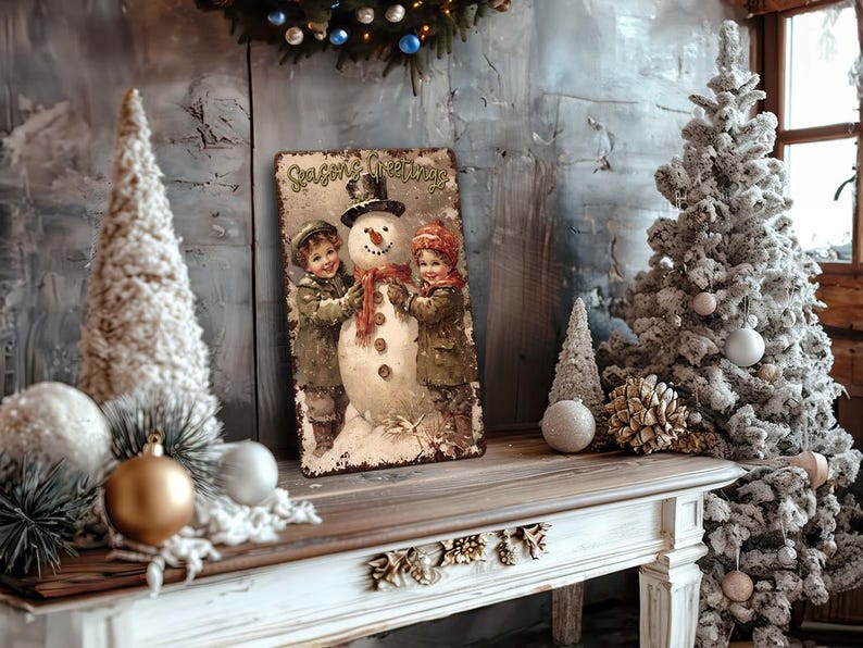 May include: A vintage-style metal sign with a snowman and two children. The sign reads "Seasons Greetings". The sign is surrounded by a white Christmas tree, a white flocked tree, and gold and white ornaments.