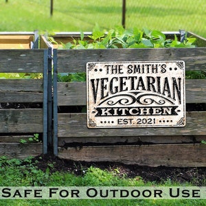 May include: A white metal sign with black text that reads "The Smith's Vegetarian Kitchen Est. 2021" is mounted on a wooden fence. The sign is in front of a garden with green plants.