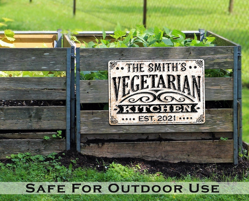 May include: A white metal sign with black text that reads "The Smith's Vegetarian Kitchen Est. 2021" is mounted on a wooden fence. The sign is in front of a garden with green plants.