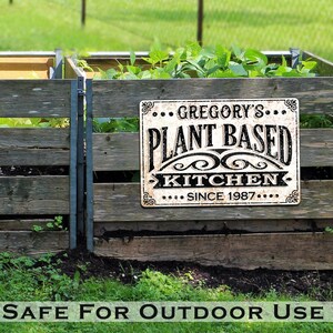 Op de afbeelding: Een verweerd wit metalen bord met zwarte letters waarop staat "Gregory's Plant Based Kitchen Since 1987". Het bord is bevestigd aan een houten hek met groene planten op de achtergrond.