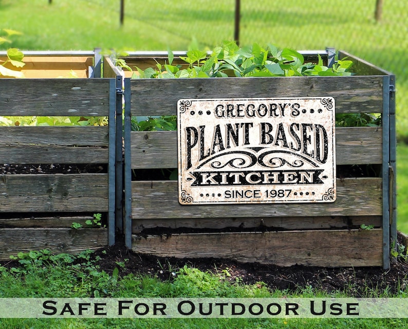 Op de afbeelding: Een verweerd wit metalen bord met zwarte letters waarop staat "Gregory's Plant Based Kitchen Since 1987". Het bord is bevestigd aan een houten hek met groene planten op de achtergrond.
