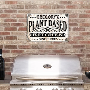 Op de afbeelding: Een wit metalen bord met zwarte tekst die "... GREGORY'S ... PLANT BASED KITCHEN ... SINCE 1987 ..." zegt. Het bord hangt aan een bakstenen muur boven een roestvrijstalen grill.