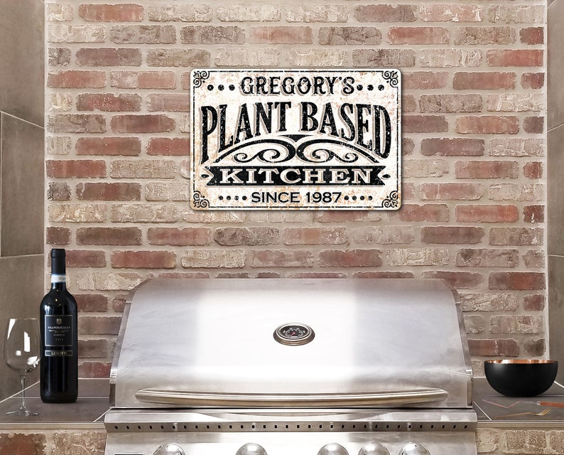 Op de afbeelding: Een wit metalen bord met zwarte tekst die "... GREGORY'S ... PLANT BASED KITCHEN ... SINCE 1987 ..." zegt. Het bord hangt aan een bakstenen muur boven een roestvrijstalen grill.