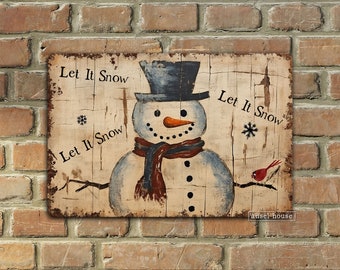 Puede incluir: Un letrero de madera rústico con un muñeco de nieve con sombrero de copa, bufanda y nariz de zanahoria. Las palabras "Let It Snow" están pintadas alrededor del muñeco de nieve. Un pájaro rojo está posado en una rama.