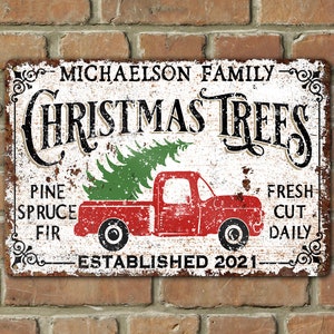 May include: A white, rusty metal sign with black text that reads "Michaelson Family Christmas Trees". A red pickup truck with a green Christmas tree in the bed is pictured on the sign. The sign also says "Pine Spruce Fir" and "Fresh Cut Daily" and "Established 2021".