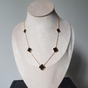 Pode incluir: Um colar dourado com pingentes pretos em forma de trevo. O colar é exibido em um manequim. Os pingentes são espaçados uniformemente ao longo da corrente, criando um design delicado. É uma joia.