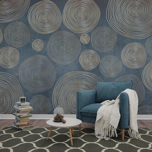 Puede incluir: Un sillón color turquesa con una manta blanca con flecos se encuentra frente a una pared con fondo azul y un patrón de espirales doradas. Una pequeña mesa blanca y una pila de libros están en primer plano. Una alfombra estampada cubre el suelo.