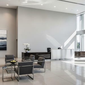 Pu&ograve; includere: Un interno di una moderna hall con poltrone grigie, tavolini in vetro e una reception bianca. Sono visibili anche un mobile in legno scuro e un dipinto con toni blu e bianchi. Il pavimento &egrave; piastrellato e la luce naturale entra da grandi finestre.