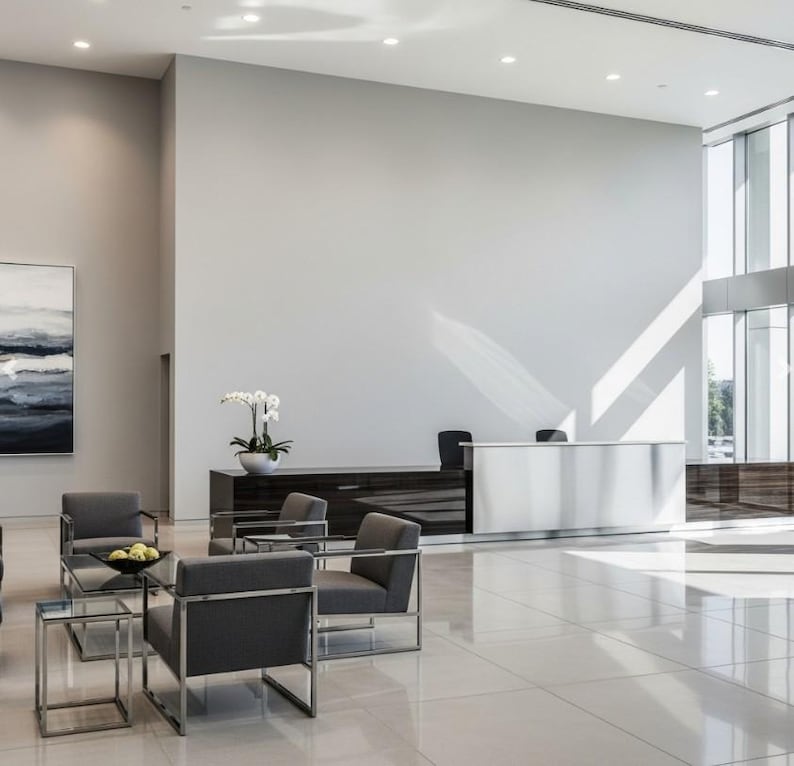 Pu&ograve; includere: Un interno di una moderna hall con poltrone grigie, tavolini in vetro e una reception bianca. Sono visibili anche un mobile in legno scuro e un dipinto con toni blu e bianchi. Il pavimento &egrave; piastrellato e la luce naturale entra da grandi finestre.
