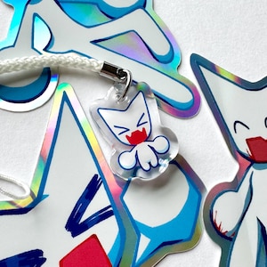 May include: A clear acrylic charm with a white cat design, red mouth, and blue accents, attached to a white cord. Other holographic stickers with similar designs are visible in the background.