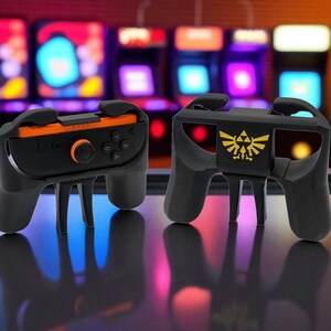 Può includere: Due impugnature nere per controller di gioco con dettagli arancioni e dorati. Un'impugnatura presenta un emblema dorato. Le impugnature sono progettate per contenere un controller di gioco. Lo sfondo mostra macchine da sala giochi.