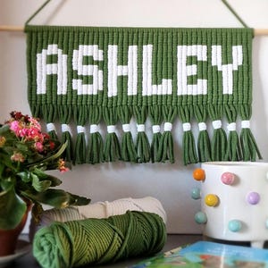 May include: A green and white macrame wall hanging with the name "ASHLEY" spelled out in white yarn. The hanging has a fringe of green and white tassels.