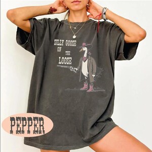 May include: A charcoal gray short-sleeve t-shirt featuring a graphic of a goose wearing a cowboy hat, trench coat, and boots while holding a revolver. The text reads SILLY GOOSE ON THE LOOSE. A small oval label in the corner displays the word PEPPER.