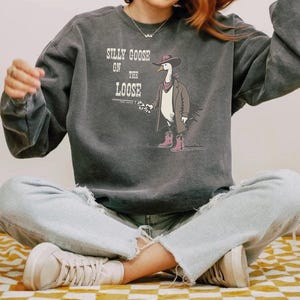 May include: A charcoal gray crewneck sweatshirt featuring a graphic of a goose wearing a brown trench coat, a pink bandana, and pink cowboy boots while holding a revolver. The text reads SILLY GOOSE ON THE LOOSE in a white, distressed font.