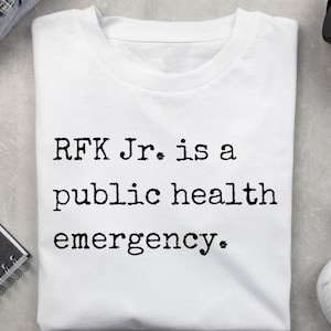 May include: White t-shirt with the text "RFK Jr. is a public health emergency" printed in black. The shirt is folded and lies on a light grey surface.