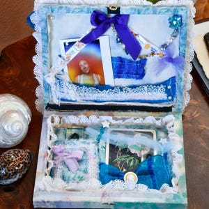 May include: Open, decorated box with a blue and white color scheme. The box is adorned with lace, ribbons, and photos. A large purple bow is tied on the top. The box is on a wooden surface.